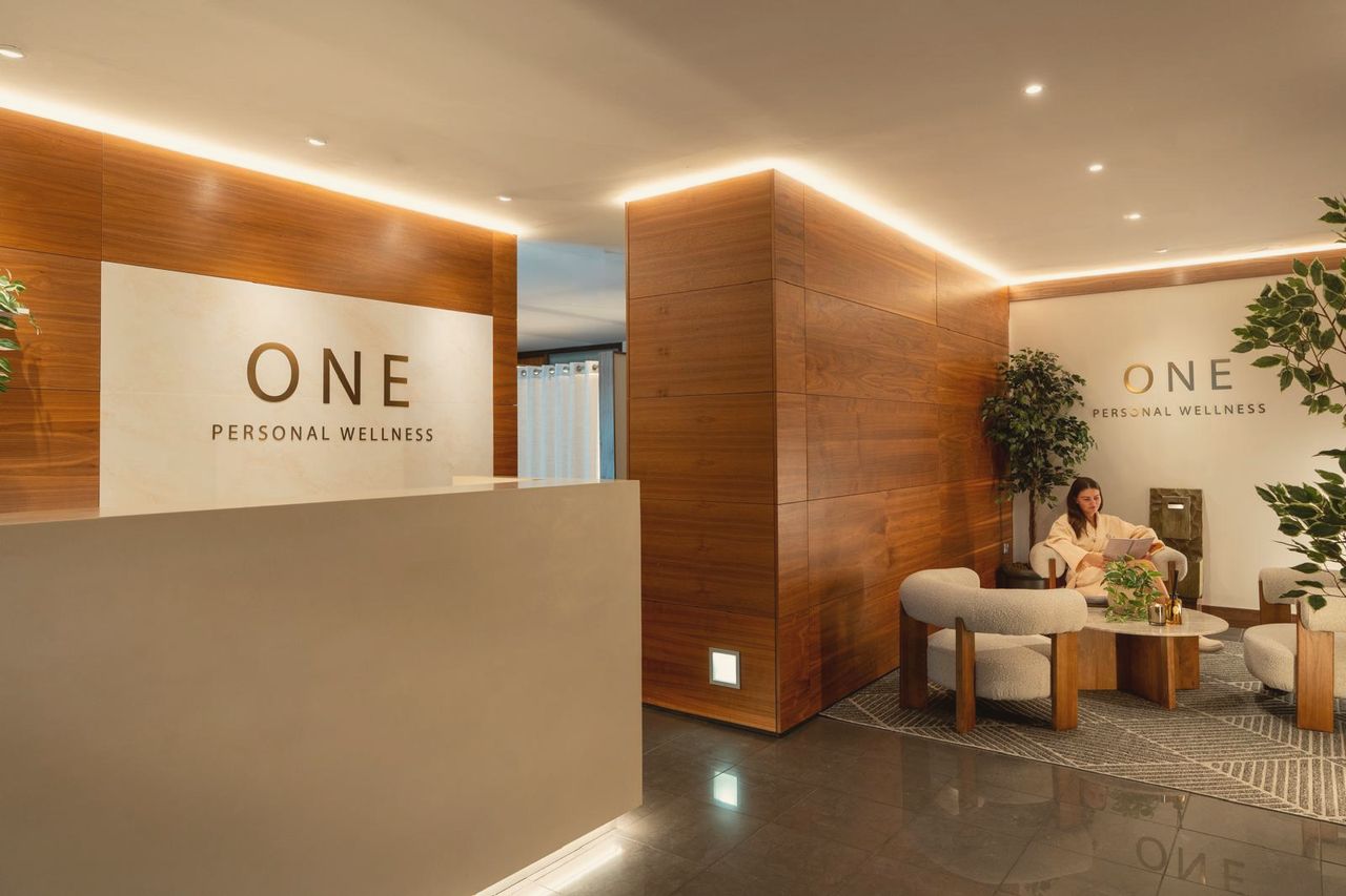 The ONE Personal Wellness reception lounge, warm lit with bouclé armchairs and a gilded wall monogram
